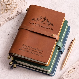 Könnte beinhalten: Ein Stapel aus drei Lederjournalen in Hellbraun, Grün und Blau. Das oberste hellbraune Journal zeigt eine Berggrafik, den Text HALEY'S JOURNAL und das Zitat Mountain and Ocean, steady as the seasons, wild as the places we love. Ein goldener Stift liegt daneben.