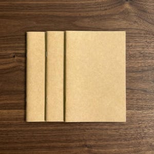 Könnte beinhalten: Drei Notizbücher aus Kraftpapier liegen auf einer dunklen Holzoberfläche. Die Notizbücher sind rechteckig mit einer einfachen, gehefteten Bindung. Das Papier hat eine natürliche, hellbraune Farbe, und die Gesamtästhetik ist minimalistisch und funktional.