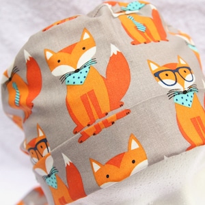 May include: A gray fabric scrub hat with a repeating pattern of orange foxes wearing blue and white bow ties. Some foxes are wearing glasses.