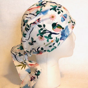 Puede incluir: Un gorro de quirófano blanco con un estampado floral colorido con pájaros y mariposas. El gorro tiene una larga corbata que se puede atar en la parte posterior del cuello. El gorro está hecho por "Scrubs & Beyond".