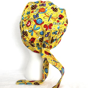 May include: Yellow fabric scrub hat with a colorful bug print. The hat has a long tie that is tied at the back of the neck. The print includes ladybugs, butterflies, bees, and dragonflies. The text "Bugs" is printed on the hat.