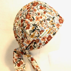 May include: A white scrub hat with a floral print in shades of orange, red, and yellow. The hat has a long tie that can be tied at the back of the neck. The hat has a small green label with the text "Green Threads" on it.
