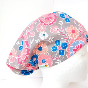 May include: A gray floral scrub cap featuring a pattern of pink, blue, and peach flowers with green leaves. The cap is made of fabric and is designed to cover the head. The flowers are in various sizes and shapes.