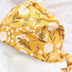 May include: A yellow fabric scrub hat with a white floral pattern. The hat has a long tie that can be tied at the back of the neck.