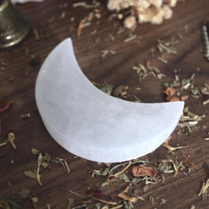 May include: A white, crescent-shaped selenite moon, resting on a dark wooden surface scattered with dried herbs and botanicals. The moon is the focal point, with a smooth, polished texture. Other objects are visible in the background.