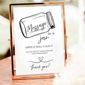 Puede incluir: Un letrero enmarcado en dorado con la frase "Message in a jar" e instrucciones para escribir, enrollar y meter un mensaje en un tarro para un aniversario. El letrero incluye un corazón y "¡gracias!"