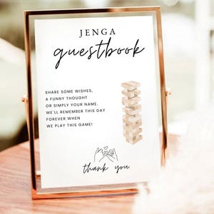 Puede incluir: Un letrero con marco dorado con las palabras "JENGA guestbook" en una elegante escritura. El letrero incluye texto que invita a los invitados a compartir deseos y un gráfico de una torre de Jenga. La parte inferior del letrero dice "thank you".