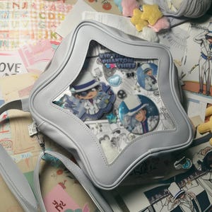 Puede incluir: Un bolso gris claro en forma de estrella con una ventana transparente que muestra insignias de personajes de anime. El bolso tiene una correa para el hombro y un cierre de cremallera. El texto "Kid the Phantom Thief" es visible.