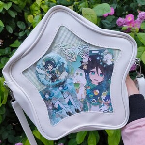 Puede incluir: Bolso ita blanco en forma de estrella con una ventana transparente que muestra arte de personajes de estilo anime, un recorte de mariposa y un pin redondo. El bolso tiene una correa para el hombro y un fondo a cuadros verde claro y blanco.