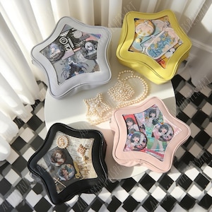 Puede incluir: Cuatro bolsas en forma de estrella en gris, amarillo, negro y rosa, cada una con una ventana transparente que muestra varios art&iacute;culos como pines y tarjetas. Tambi&eacute;n hay un collar de perlas y joyas en forma de estrella. Las bolsas est&aacute;n sobre una superficie blanca con un suelo a cuadros blanco y negro.