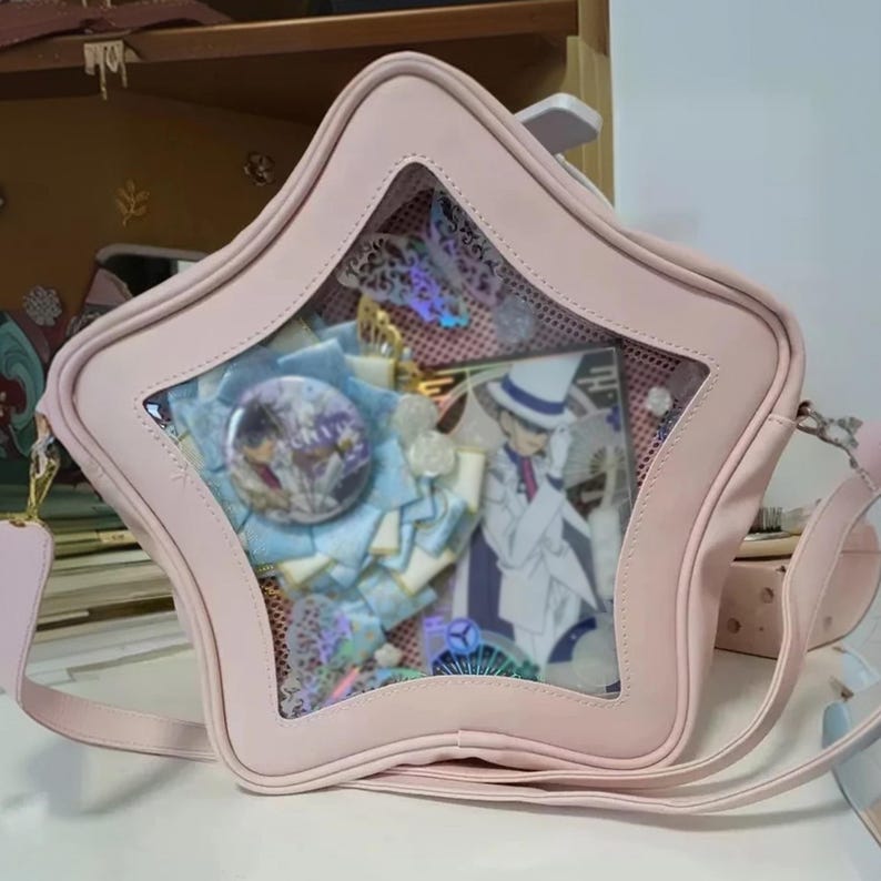 Puede incluir: Un bolso ita rosa en forma de estrella con una ventana transparente, que muestra accesorios tem&aacute;ticos de anime. El bolso tiene una correa para el hombro y un cierre de cremallera. En el interior, hay insignias, cintas y una tarjeta de personaje.