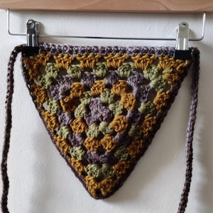 May include: A triangular crocheted scarf with brown, green, and yellow yarn. The scarf has long brown ties.