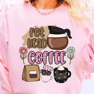 Può includere: Felpa rosa con un design grafico di una caffettiera, tazze da caffè e chicchi di caffè. Il testo sulla felpa dice "Pot Head Coffee".