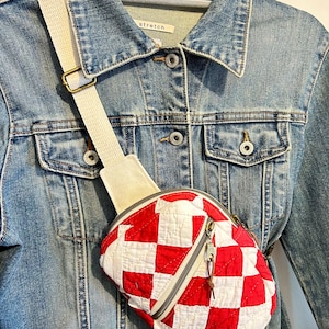 Peut inclure: Un sac banane matelassé à motif damier rouge et blanc, doté d'une poche zippée en diagonale et d'une large sangle réglable couleur crème, présenté sur une veste en jean bleue avec des boutons métalliques et une étiquette indiquant stretch.