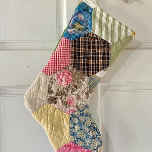 May include: A quilted Christmas stocking constructed from various fabric patches in a hexagonal pattern. The patchwork features diverse colors and prints, including red and white gingham, brown plaid, floral patterns, and solid yellow and pink.