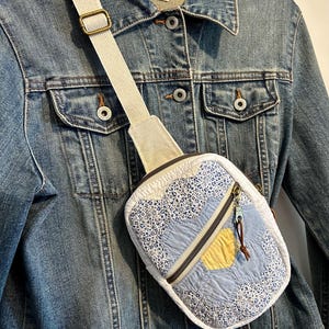 Peut inclure: Un sac bandoulière matelassé présentant un motif patchwork floral et uni dans des tons bleu clair et jaune. Le sac est doté d'une sangle en toile blanche réglable avec une boucle en métal doré et d'une fermeture éclair avec une tirette en cuir.