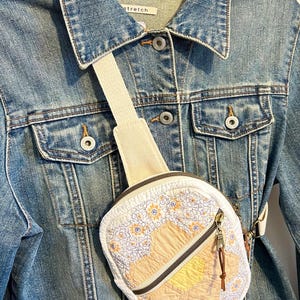 May include: A blue denim jacket with a collar and metal buttons, featuring a small, round, quilted crossbody bag. The bag has a white background with a floral pattern, a central yellow and beige hexagonal patch, and a dark metal zipper closure.