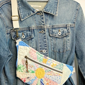May include: A quilted bum bag featuring a sunburst pattern made of various pastel-coloured floral and checkered fabric patches. The bag has a horizontal zippered pocket with a brown leather pull tab and an adjustable cream-coloured canvas strap.