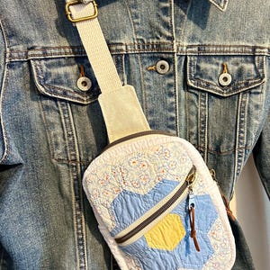 May include: A quilted crossbody bag featuring a patchwork design of light blue, yellow, and white floral fabric. The bag has a beige canvas strap with a metal buckle and a zippered front pocket with a brown leather pull tab, worn over a blue denim jacket.