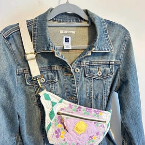May include: A denim jacket with a white and floral fanny pack. The fanny pack has a zipper closure and a white strap with a gold-colored buckle. The fanny pack is decorated with a large purple flower and green diamond shapes.