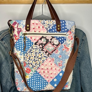 May include: A quilted tote bag featuring a patchwork design of various floral and polka dot patterns in blue, pink, and white. The bag includes dark brown leather handles, a brown fabric shoulder strap, and a zippered front pocket with brass hardware.