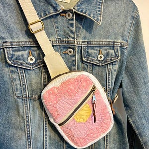 Peut inclure: Un sac bandoulière matelassé avec un motif patchwork hexagonal rose et blanc et un accent hexagonal jaune. Le sac possède une sangle en toile blanche, une fermeture éclair en métal argenté et une tirette en cuir marron, porté sur une veste en jean.