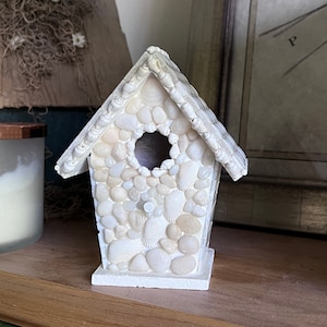 May include: A white birdhouse crafted from seashells and small white stones. The birdhouse features a small opening for a bird to enter.