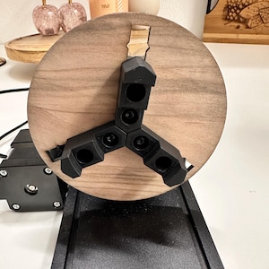 May include: A close-up of a wood turning lathe with a wooden disc attached to the chuck. The chuck has three black arms with a series of holes in each arm.