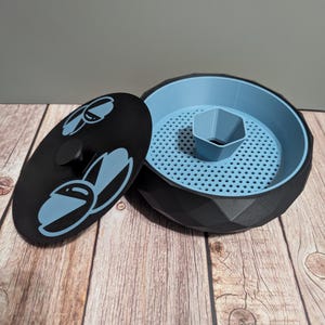 Puede incluir: Un recipiente negro y azul con tapa. El recipiente tiene un diseño geométrico y un interior azul claro. La tapa es negra con un diseño azul. El recipiente está sobre una superficie de madera.