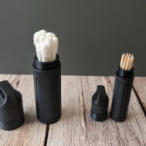 Könnte beinhalten: Schwarze, strukturierte zylindrische Behälter mit Schraubdeckeln. Ein Behälter enthält weiße Wattestäbchen, ein anderer Zahnstocher. Die Behälter stehen auf einer Holzoberfläche mit dunklem grauem Hintergrund.