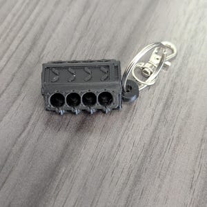 Peut inclure: Un porte-clés en plastique noir en forme de bloc moteur de voiture. Le porte-clés a un anneau en métal argenté et un fermoir en métal argenté.