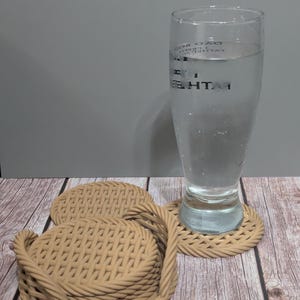 May include: A clear glass filled with water sits on a beige woven coaster. Two additional coasters are next to it. The coasters are made of a woven material and are tan in color. The background is a wooden surface.