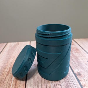 May include: Teal-colored cylindrical container with a matching lid. The container has a textured geometric pattern and a threaded opening. The lid is oval-shaped with a series of oval cutouts. The container is open, revealing the interior.