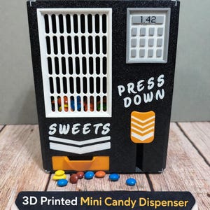 Könnte beinhalten: Ein schwarzer, 3D-gedruckter Mini-Bonbon-Spender mit einem weißen Gitter, einem grauen Tastenfeld und den Wörtern "SWEETS" und "PRESS DOWN". Der Spender hat einen orangefarbenen Ausgabeschlitz und ist mit bunten Süßigkeiten gefüllt. Der Text "3D Printed Mini Candy Dispenser" ist ebenfalls sichtbar.