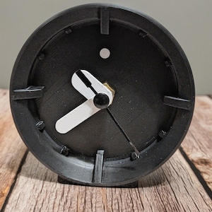 May include: A black analog clock with white hands and a black face. The clock has a round shape and is sitting on a wooden surface. The clock's hands point to approximately 2:40.