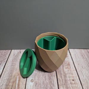 Puede incluir: Un recipiente impreso en 3D con un diseño geométrico. El exterior es de color beige y el interior es verde. La tapa, también verde, se coloca junto al recipiente. El recipiente está abierto, revelando el interior.