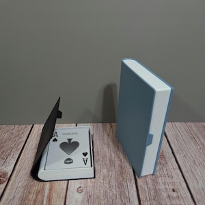 Peut inclure: Un jeu de cartes avec l'As de Pique visible, présenté dans un étui en forme de livre. Une boîte bleue fermée en forme de livre est également présente. Le design suggère un thème de mystère et d'éléments cachés.