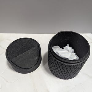 May include: A black, textured, small trash can with a lid. The lid is round and flat, with a speckled surface. The can's body is cylindrical with a diamond pattern. The can is open, revealing crumpled white paper inside.