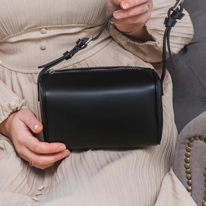 May include: A black leather crossbody bag with a cylindrical shape and a zip closure. The bag has a long, adjustable strap. The bag is held by a person wearing a beige dress. The bag's design is minimalist.