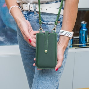 Puede incluir: Una funda de tel&eacute;fono de cuero verde oscuro con una correa verde a juego. La funda tiene un cierre de bot&oacute;n dorado y la sostiene una persona que lleva vaqueros azules y un cintur&oacute;n blanco. La correa est&aacute; sujeta con cierres dorados.