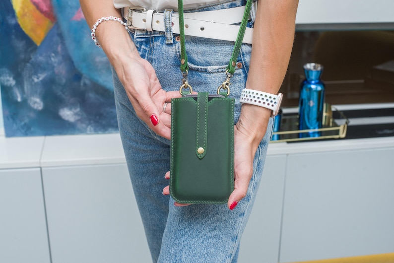 Puede incluir: Una funda de tel&eacute;fono de cuero verde oscuro con una correa verde a juego. La funda tiene un cierre de bot&oacute;n dorado y la sostiene una persona que lleva vaqueros azules y un cintur&oacute;n blanco. La correa est&aacute; sujeta con cierres dorados.