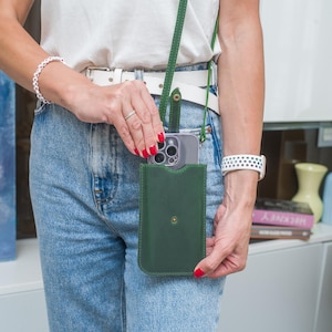 Puede incluir: Una funda de tel&eacute;fono de cuero verde oscuro con una correa a juego. La funda tiene un dise&ntilde;o sencillo con un solo cierre de bot&oacute;n dorado. La funda del tel&eacute;fono la sostiene una persona que lleva una camisa blanca y vaqueros azules.