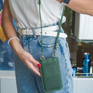 Puede incluir: Bolso bandolera verde de cuero para tel&eacute;fono con una correa larga verde. El bolso tiene ranuras para tarjetas y herrajes dorados. La persona lleva una camisa blanca, vaqueros azules y un cintur&oacute;n blanco.
