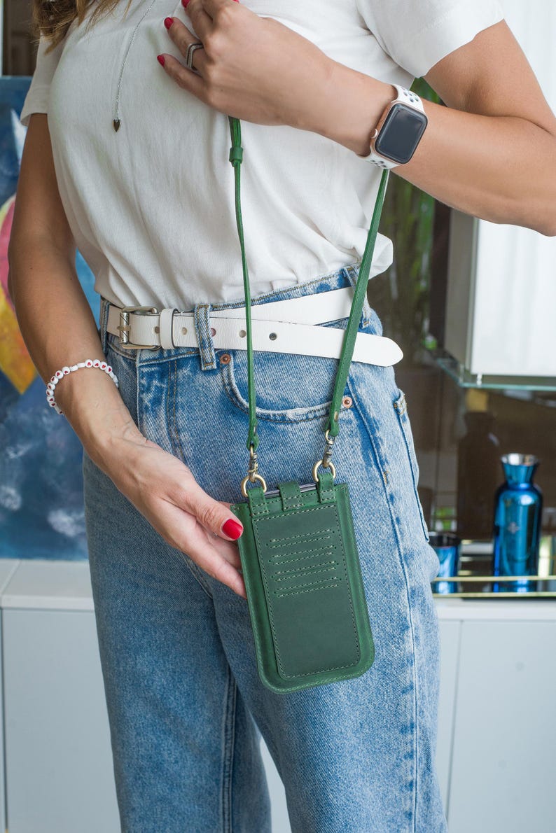 Puede incluir: Bolso bandolera verde de cuero para tel&eacute;fono con una correa larga verde. El bolso tiene ranuras para tarjetas y herrajes dorados. La persona lleva una camisa blanca, vaqueros azules y un cintur&oacute;n blanco.