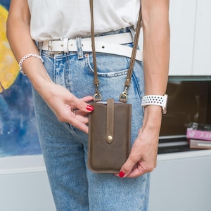 Könnte beinhalten: Eine braune Leder-Umhängetasche für ein Telefon mit langem Riemen. Die Tasche hat eine vertikale Klappe mit einem goldfarbenen Knopfverschluss. Die Person trägt einen weißen Gürtel, blaue Jeans und ein weißes Hemd. Eine weiße Uhr ist am Handgelenk.