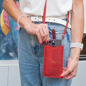 Puede incluir: Funda de tel&eacute;fono cruzada de cuero rojo con una correa a juego. La funda tiene un cierre a presi&oacute;n y contiene un tel&eacute;fono inteligente. La persona lleva un cintur&oacute;n blanco, vaqueros azules y una camisa blanca. Tambi&eacute;n se ven una pulsera y un reloj blancos.