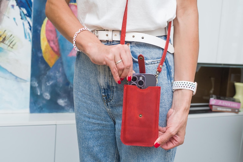 Puede incluir: Funda de tel&eacute;fono cruzada de cuero rojo con una correa a juego. La funda tiene un cierre a presi&oacute;n y contiene un tel&eacute;fono inteligente. La persona lleva un cintur&oacute;n blanco, vaqueros azules y una camisa blanca. Tambi&eacute;n se ven una pulsera y un reloj blancos.