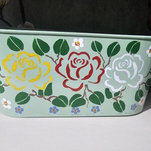 May include: A rectangular, pale green metal planter with two handles. It features yellow, red, and white rose designs, green leaves, and small white and blue flowers. The planter is on a white surface.