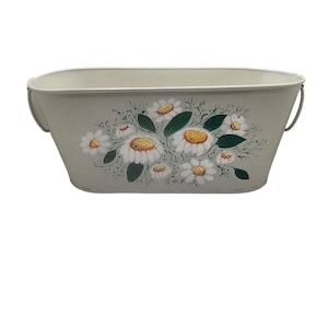 Peut inclure: Une jardinière rectangulaire en métal gris clair ornée d'un motif floral de marguerites. Les marguerites ont des pétales blancs et des centres jaunes, avec des feuilles et des tiges vertes. La jardinière a de petites poignées en métal de chaque côté.