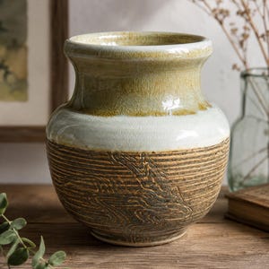 Könnte beinhalten: Eine Keramikvase mit einem breiten, ausgestellten Rand und einem bauchigen Körper. Der obere Teil zeigt eine glänzende, rissige Glasur in Olivgrün und Cremetönen, während der untere Bereich eine texturierte, dunkelbraune Oberfläche mit Rillen aufweist.